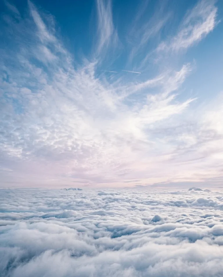 Serene pastel sunrise sky with soft wispy clouds drifting above a peaceful sea of clouds.